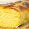 Pão brioche em forma de trança, grande e dourado, com um aspeto brilhante, pousado sobre uma tábua de madeira rústica.