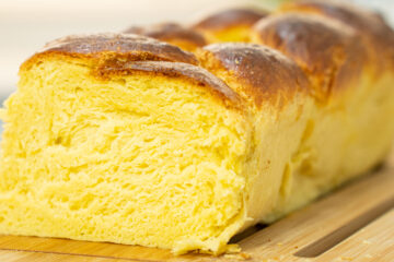 Pão brioche em forma de trança, grande e dourado, com um aspeto brilhante, pousado sobre uma tábua de madeira rústica.
