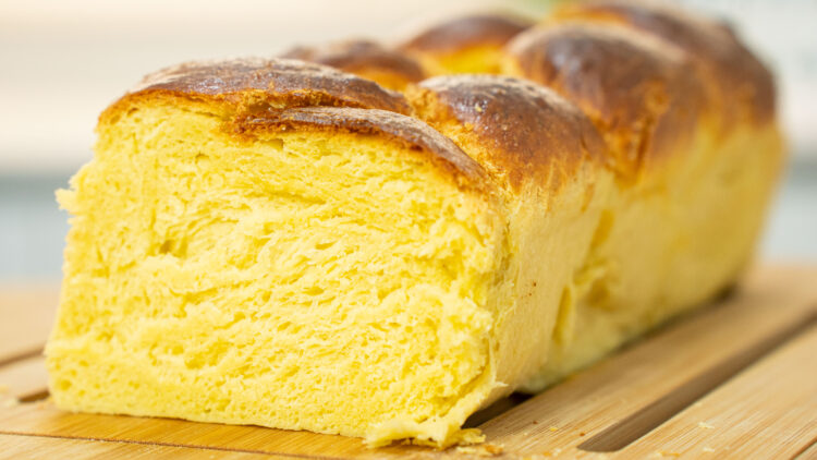 Pão brioche em forma de trança, grande e dourado, com um aspeto brilhante, pousado sobre uma tábua de madeira rústica.