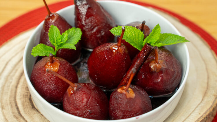 Pera rocha cozida e de pé num prato branco, banhada por uma calda de vinho espessa e brilhante, decorada com pau de canela e folha de hortelã.