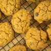 Várias cookies douradas e redondas com pepitas de chocolate negro salientes, dispostas sobre uma grelha de arrefecimento de metal.
