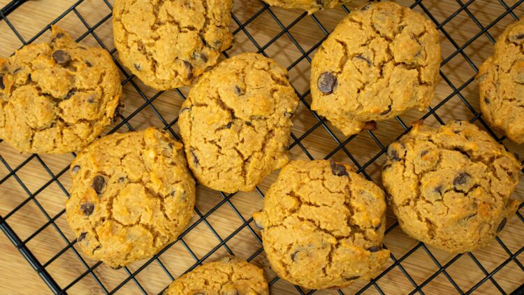 Várias cookies douradas e redondas com pepitas de chocolate negro salientes, dispostas sobre uma grelha de arrefecimento de metal.