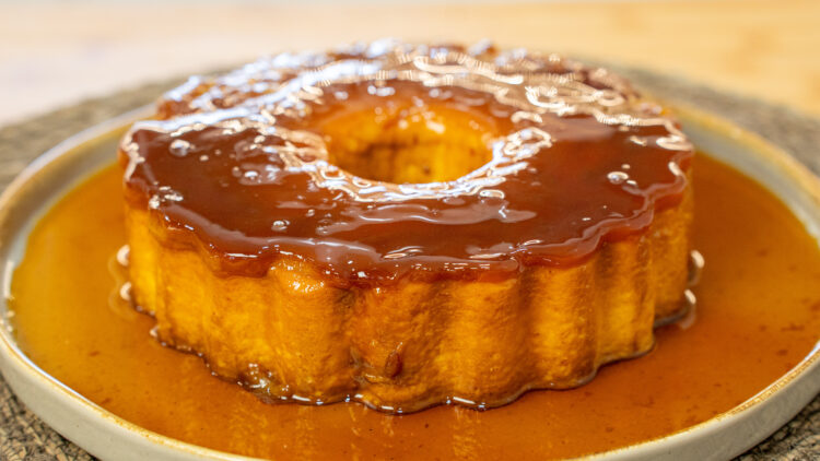 Pudim redondo e liso com uma cor amarela suave, banhado por uma calda de caramelo dourada e brilhante, servido num prato raso.