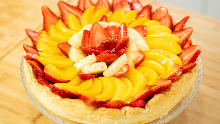 Tarte redonda assente num prato com pé, com uma base de massa folhada rústica e coberta de forma abundante e em espiral por fatias brilhantes de morangos vermelhos, pêssegos laranjas e ananás amarelo.