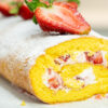 Uma torta cilíndrica de pão de ló, recheada com natas brancas e pedaços vermelhos de morango, polvilhada generosamente com açúcar em pó e decorada com morangos inteiros cortados ao meio no topo.