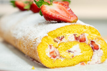 Uma torta cilíndrica de pão de ló, recheada com natas brancas e pedaços vermelhos de morango, polvilhada generosamente com açúcar em pó e decorada com morangos inteiros cortados ao meio no topo.