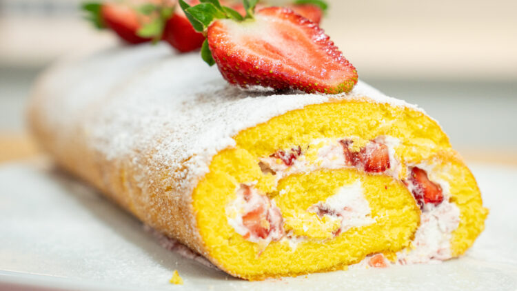Uma torta cilíndrica de pão de ló, recheada com natas brancas e pedaços vermelhos de morango, polvilhada generosamente com açúcar em pó e decorada com morangos inteiros cortados ao meio no topo.