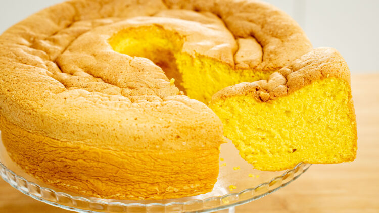 Um Pão de Ló grande e dourado, com um aspeto rústico e fofinho, servido num prato redondo de cor clara. O bolo tem a clássica forma redonda com buraco no meio.