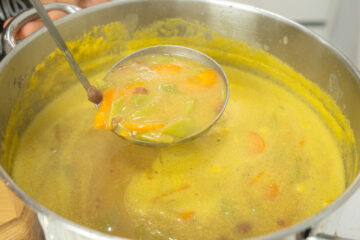 Prato fundo de barro com uma sopa cremosa de cor acastanhada, repleta de feijão verde cortado, meias-luas de cenoura laranja e feijões manteiga inteiros.