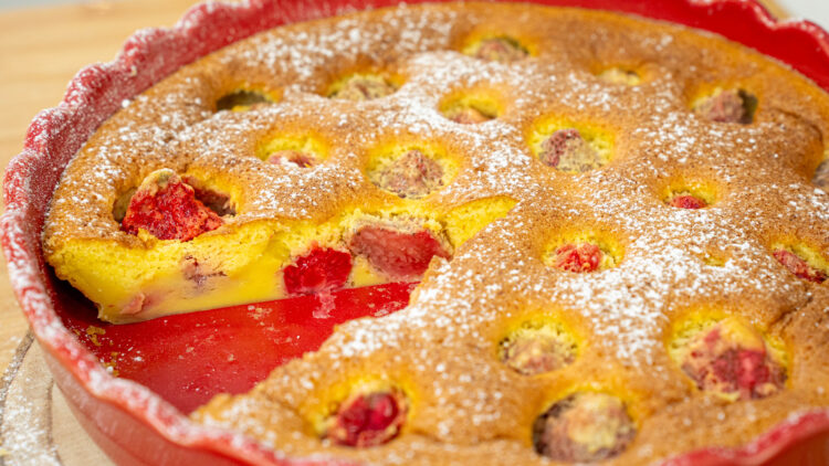 Tarte rústica cozida numa forma de barro redonda, generosamente polvilhada com açúcar em pó branco, com ligeiros relevos avermelhados de morangos no interior da massa.