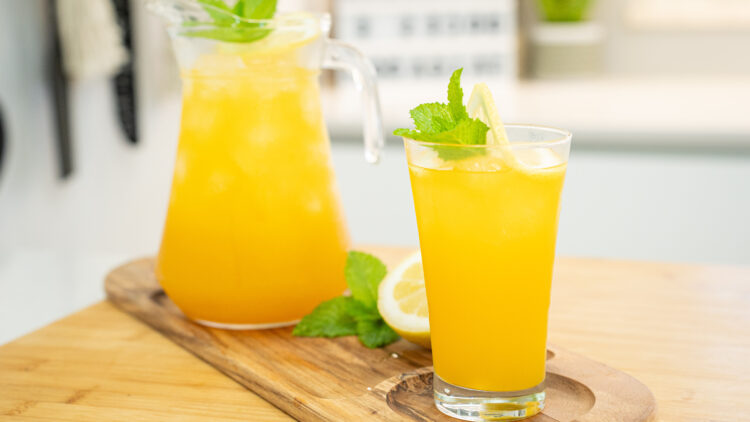 Jarra de vidro de 1 litro, suada devido ao gelo, cheia com uma bebida amarelo-alaranjada, decorada com fatias de limão e folhas verdes de hortelã fresca no topo.