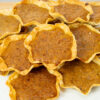 Várias queijadas pequenas, douradas e redondas, com uma base de massa visivelmente fina e estaladiça, revelando um recheio cozido com pedaços de noz. Servidas num prato claro e elegante.