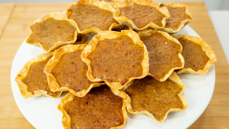 Várias queijadas pequenas, douradas e redondas, com uma base de massa visivelmente fina e estaladiça, revelando um recheio cozido com pedaços de noz. Servidas num prato claro e elegante.