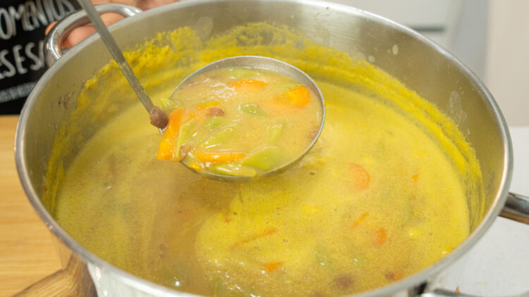 Prato fundo de barro com uma sopa cremosa de cor acastanhada, repleta de feijão verde cortado, meias-luas de cenoura laranja e feijões manteiga inteiros.