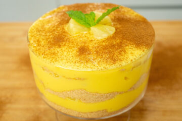 Taça redonda de vidro transparente com uma sobremesa de colher em camadas, mostrando linhas alternadas de creme amarelo pálido, bolacha de canela triturada e pedaços de ananás. O topo está polvilhado artisticamente com canela em pó e decorado no centro com três cubos frescos de ananás.