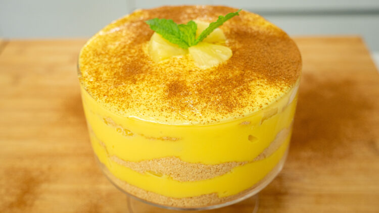 Taça redonda de vidro transparente com uma sobremesa de colher em camadas, mostrando linhas alternadas de creme amarelo pálido, bolacha de canela triturada e pedaços de ananás. O topo está polvilhado artisticamente com canela em pó e decorado no centro com três cubos frescos de ananás.