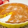 Travessa rústica redonda de barro com uma tarte de maçã tipo flã no interior, exibindo as fatias de maçã caramelizadas na superfície e um ligeiro polvilhado branco de açúcar em pó e canela.