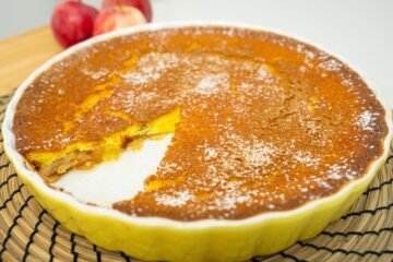 Travessa rústica redonda de barro com uma tarte de maçã tipo flã no interior, exibindo as fatias de maçã caramelizadas na superfície e um ligeiro polvilhado branco de açúcar em pó e canela.