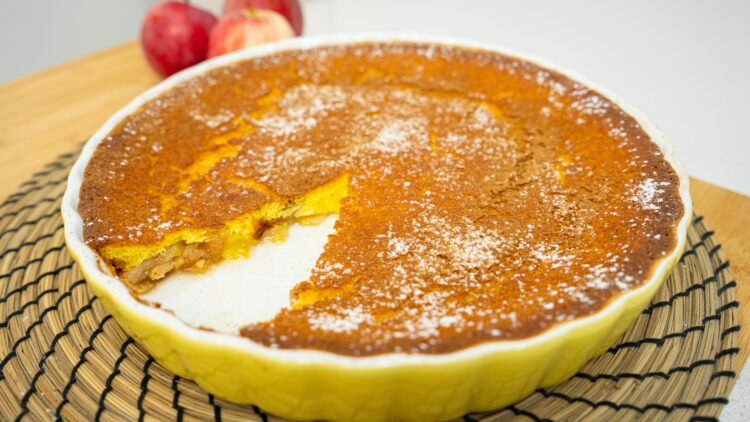 Travessa rústica redonda de barro com uma tarte de maçã tipo flã no interior, exibindo as fatias de maçã caramelizadas na superfície e um ligeiro polvilhado branco de açúcar em pó e canela.