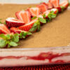 Travessa de vidro quadrada com um tiramisu cremoso, polvilhado finamente com cacau em pó escuro e decorado simetricamente com metades de morangos vermelhos brilhantes com folha verde e folhas de hortelã fresca