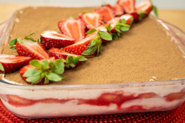 Travessa de vidro quadrada com um tiramisu cremoso, polvilhado finamente com cacau em pó escuro e decorado simetricamente com metades de morangos vermelhos brilhantes com folha verde e folhas de hortelã fresca