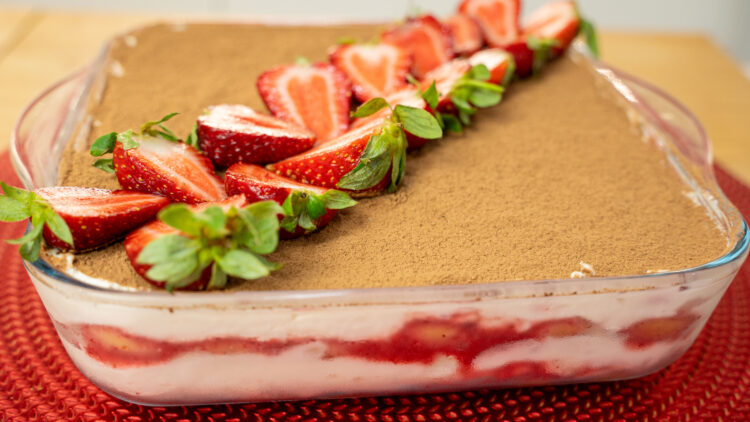 Travessa de vidro quadrada com um tiramisu cremoso, polvilhado finamente com cacau em pó escuro e decorado simetricamente com metades de morangos vermelhos brilhantes com folha verde e folhas de hortelã fresca