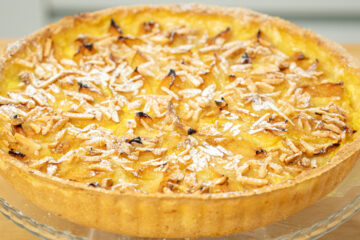 Tarte redonda rústica de 27 centímetros disposta num prato de servir, com rebordos de massa dourada, cobertura densa de pedaços de ameixa amarela cozida e lascas de amêndoa palitada tostada, salpicada com açúcar em pó.