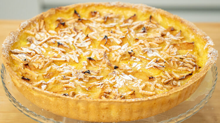 Tarte redonda rústica de 27 centímetros disposta num prato de servir, com rebordos de massa dourada, cobertura densa de pedaços de ameixa amarela cozida e lascas de amêndoa palitada tostada, salpicada com açúcar em pó.