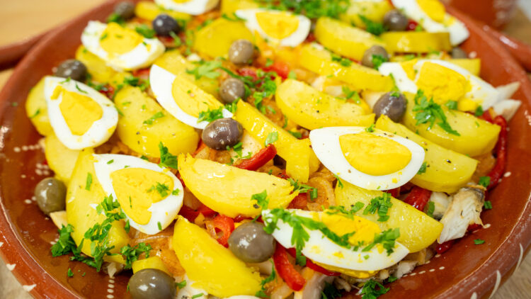 Uma rústica travessa de barro retangular, cheia de um prato gratinado com camadas de batata e peixe, decorada no topo com metades de ovo cozido, azeitonas pretas e salsa picada.