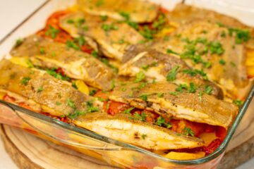 Travessa de vidro transparente com um suculento assado de forno, exibindo pedaços de peixe branco (solha) cozinhados sobre uma cama rica de rodelas finas de batata, cebola, tiras de pimento verde e fatias de tomate, tudo polvilhado com salsa fresca picada.