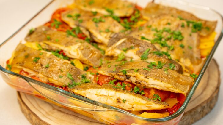 Travessa de vidro transparente com um suculento assado de forno, exibindo pedaços de peixe branco (solha) cozinhados sobre uma cama rica de rodelas finas de batata, cebola, tiras de pimento verde e fatias de tomate, tudo polvilhado com salsa fresca picada.