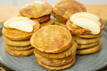 Uma pilha apetitosa de panquecas redondas e douradas, servidas num prato claro, decoradas com um fio brilhante de mel a escorrer pelas laterais e acompanhadas por finas fatias de maçã fresca.