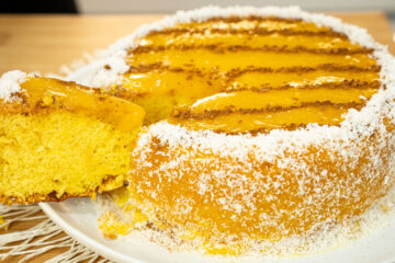 Bolo alto e redondo coberto de creme amarelo brilhante (doce de ovos), decorado no topo com linhas paralelas de canela em pó, e com toda a base contornada por flocos brancos de coco ralado.