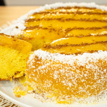 Bolo alto e redondo coberto de creme amarelo brilhante (doce de ovos), decorado no topo com linhas paralelas de canela em pó, e com toda a base contornada por flocos brancos de coco ralado.