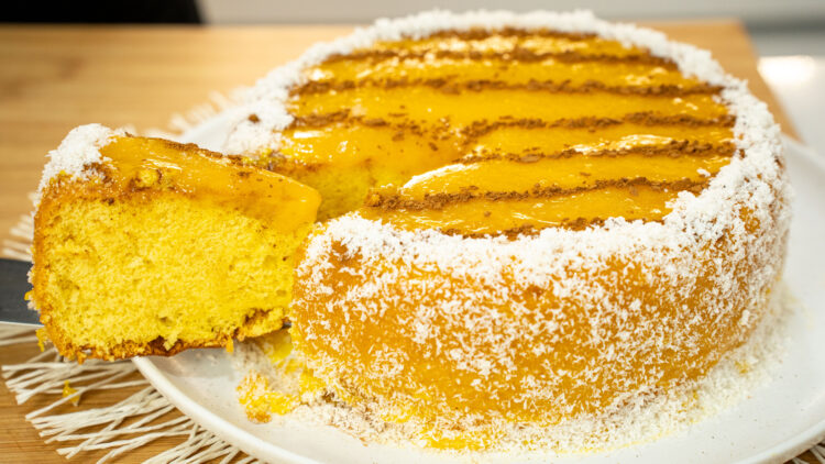 Bolo alto e redondo coberto de creme amarelo brilhante (doce de ovos), decorado no topo com linhas paralelas de canela em pó, e com toda a base contornada por flocos brancos de coco ralado.