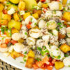 Travessa farta e colorida dividida em secções: chocos grelhados com marcas da chapa, batatas cozidas brancas e uma salada vibrante de tomate e pepino, coberta generosamente com cubos crocantes e dourados de broa torrada e polvilhada com salsa fresca.