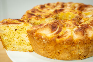 Bolo de buraco dourado e alto, servido num prato branco de pé, com o topo decorado por fatias finas de pera assada dispostas em espiral e um polvilhado rústico de canela.