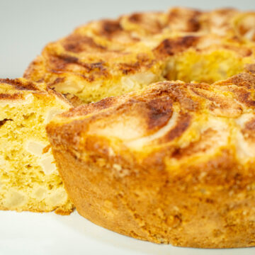 Bolo de buraco dourado e alto, servido num prato branco de pé, com o topo decorado por fatias finas de pera assada dispostas em espiral e um polvilhado rústico de canela.