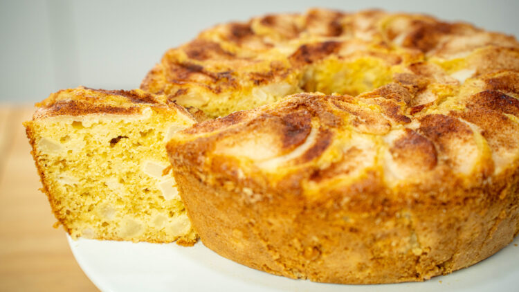 Bolo de buraco dourado e alto, servido num prato branco de pé, com o topo decorado por fatias finas de pera assada dispostas em espiral e um polvilhado rústico de canela.