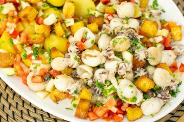 Travessa farta e colorida dividida em secções: chocos grelhados com marcas da chapa, batatas cozidas brancas e uma salada vibrante de tomate e pepino, coberta generosamente com cubos crocantes e dourados de broa torrada e polvilhada com salsa fresca.