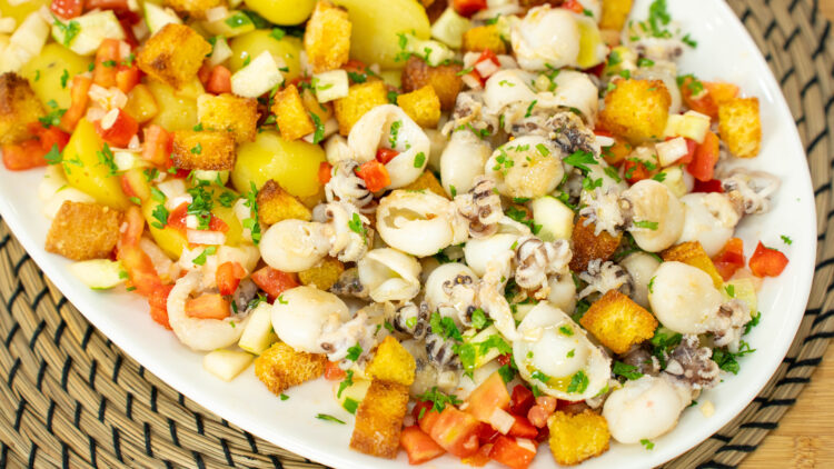 Travessa farta e colorida dividida em secções: chocos grelhados com marcas da chapa, batatas cozidas brancas e uma salada vibrante de tomate e pepino, coberta generosamente com cubos crocantes e dourados de broa torrada e polvilhada com salsa fresca.