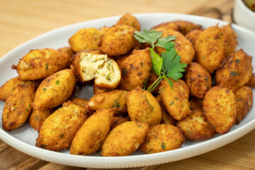 Uma travessa branca repleta de pastéis de pescada perfeitamente fritos, com uma cor dourada uniforme e formato tradicional alongado, repousando sobre uma cama de papel absorvente.