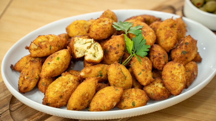 Uma travessa branca repleta de pastéis de pescada perfeitamente fritos, com uma cor dourada uniforme e formato tradicional alongado, repousando sobre uma cama de papel absorvente.