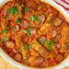 Travessa rústica de barro repleta de arroz assado com um tom dourado escuro, guarnecido no topo com pedaços de entrecosto de porco suculentos, rodelas de chouriço, tiras de pimento vermelho e polvilhado com salsa fresca picada.