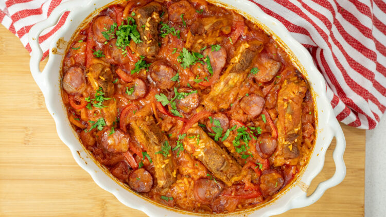 Travessa rústica de barro repleta de arroz assado com um tom dourado escuro, guarnecido no topo com pedaços de entrecosto de porco suculentos, rodelas de chouriço, tiras de pimento vermelho e polvilhado com salsa fresca picada.