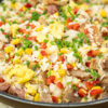 Frigideira larga preta cheia de arroz salteado soltinho, misturado com pedaços dourados de carne grelhada, grãos amarelos de milho, cubos de pimento vermelho e pedaços de ovo mexido, polvilhado com cebolinho picado.