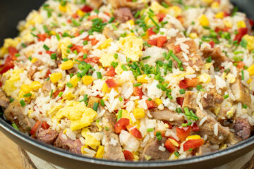 Frigideira larga preta cheia de arroz salteado soltinho, misturado com pedaços dourados de carne grelhada, grãos amarelos de milho, cubos de pimento vermelho e pedaços de ovo mexido, polvilhado com cebolinho picado.