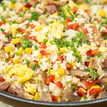 Frigideira larga preta cheia de arroz salteado soltinho, misturado com pedaços dourados de carne grelhada, grãos amarelos de milho, cubos de pimento vermelho e pedaços de ovo mexido, polvilhado com cebolinho picado.