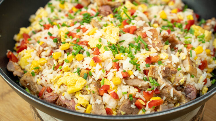 Frigideira larga preta cheia de arroz salteado soltinho, misturado com pedaços dourados de carne grelhada, grãos amarelos de milho, cubos de pimento vermelho e pedaços de ovo mexido, polvilhado com cebolinho picado.