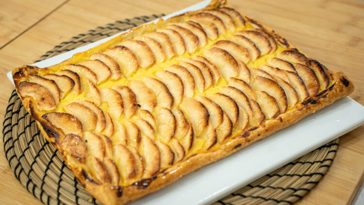 Tarte retangular de massa folhada, exibindo um rebordo alto, dourado e estaladiço, com um interior recheado de creme amarelo coberto por finas meias-luas de maçã assada e brilhante (pincelada com mel).